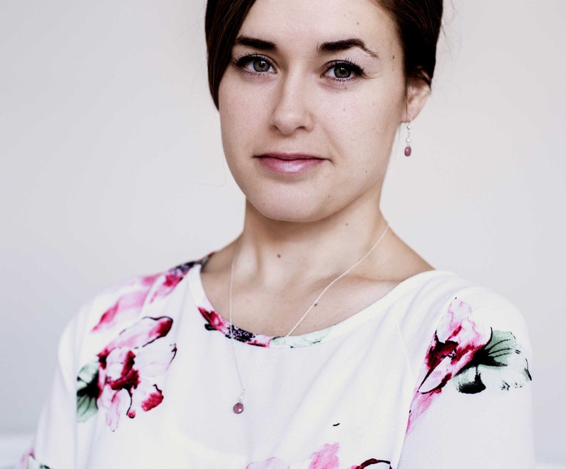Puede incluir: Una mujer con cabello casta&ntilde;o que lleva una blusa blanca con estampado floral. Lleva un collar de plata con un colgante de piedra rosa y pendientes de plata con detalles de piedra rosa.