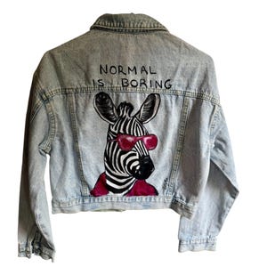 Op de afbeelding: Een lichtblauw acid-wash spijkerjack met een handgeschilderde illustratie op de achterkant van een zebra met een roze zonnebril en een roze sjaal. De tekst "NORMAL IS BORING" is in het zwart boven de zebra-illustratie op de rug gedrukt.