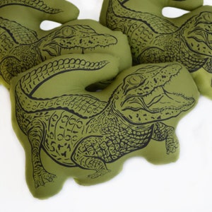 May include: Three green fabric pillows with black alligator designs. The pillows are shaped like alligators and have a textured fabric.