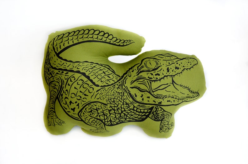 May include: A green pillow shaped like an alligator with a black outline. The alligator's mouth is open and its teeth are visible.