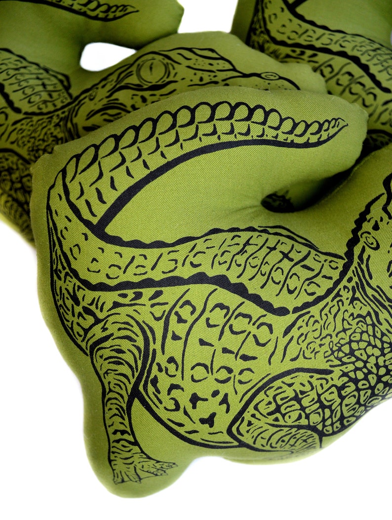May include: Three green fabric alligator pillows with black alligator print designs. The pillows are shaped like alligators and have a textured fabric.