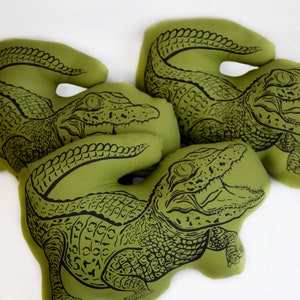 May include: Three green fabric pillows with black alligator designs. The alligators are in different poses, with one facing forward, one facing to the side, and one facing backward.