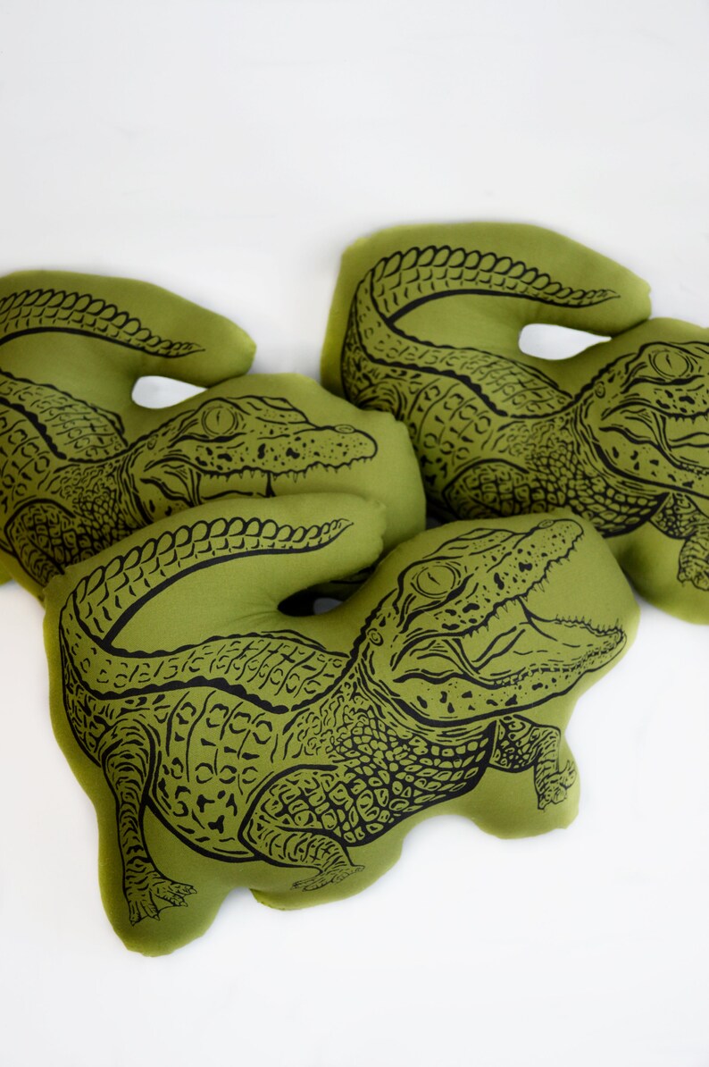 May include: Three green fabric pillows with black alligator designs. The alligators are in different poses, with one facing forward, one facing to the side, and one facing backward.