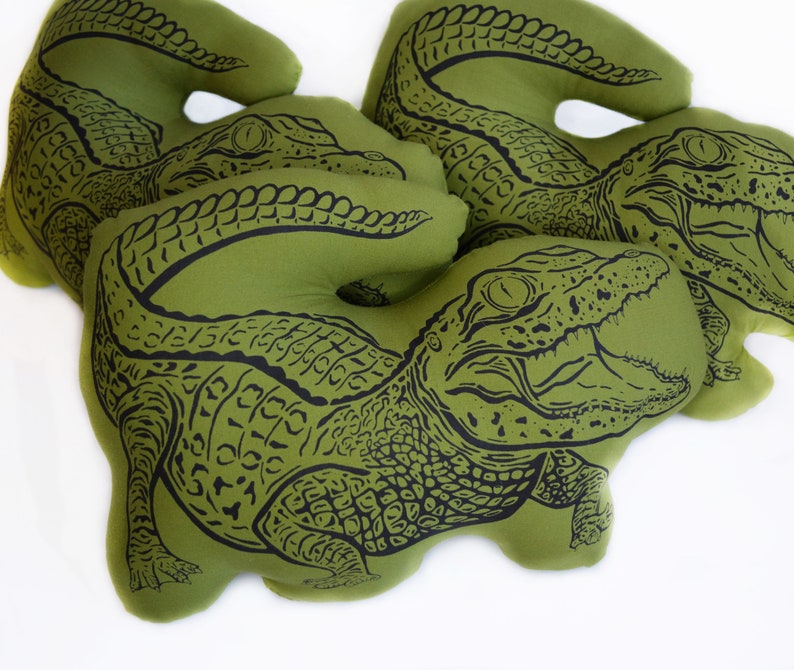 May include: Three green fabric pillows with black alligator designs. The pillows are shaped like alligators with their mouths open.