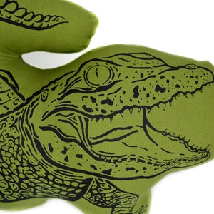 May include: Green fabric pillow with a black and white illustration of an alligator with its mouth open. The alligator has a scaly texture and a detailed eye.