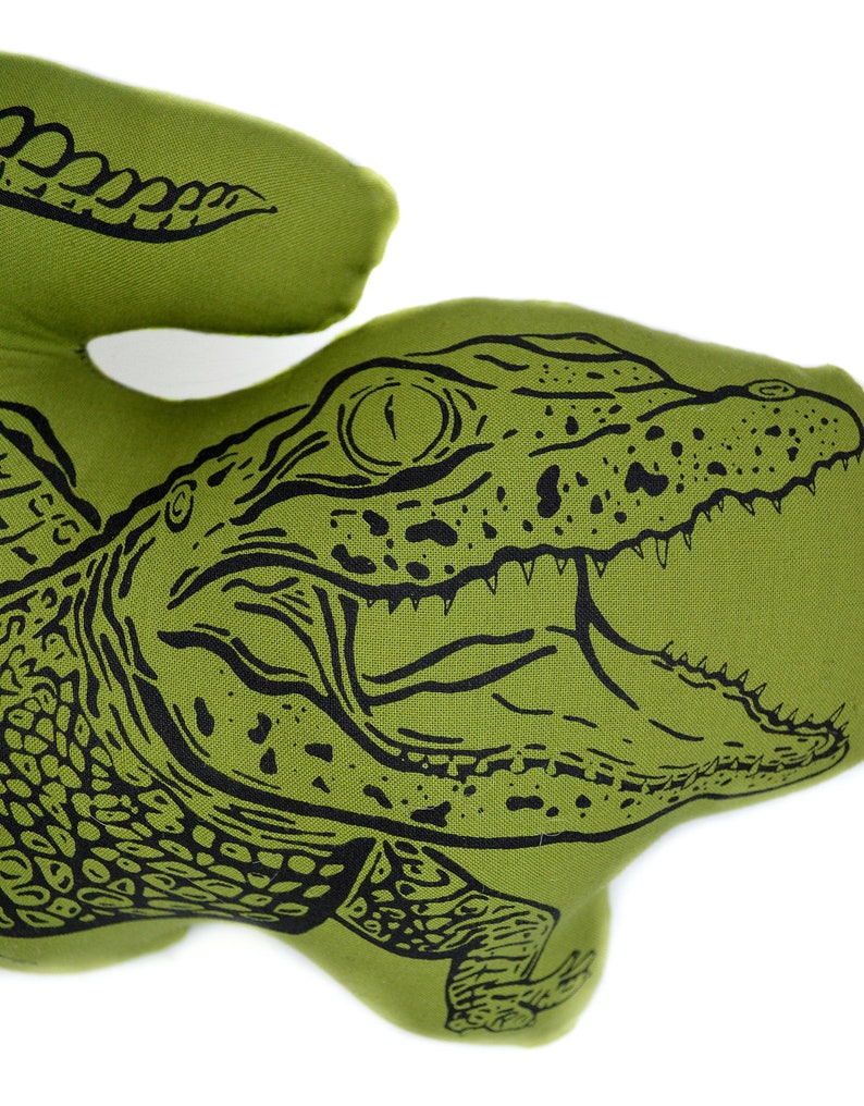 May include: Green fabric pillow with a black and white illustration of an alligator with its mouth open. The alligator has a scaly texture and a detailed eye.
