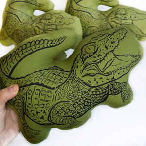May include: Green plush alligator toy with black embroidered details. The alligator has a wide open mouth and a textured body.