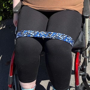 Op de afbeelding: Een persoon in een rolstoel draagt een bruine trui, zwarte leggings en een blauw-witte weerstandsband om de dijen. De persoon glimlacht en draagt een bril. De achtergrond is een groene haag.