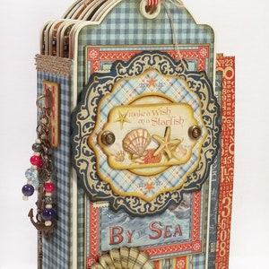May include: A multi-layered paper craft album with a nautical theme, featuring a blue gingham pattern, a central plaque reading "make a wish on a Starfish" with seashell and starfish motifs, and a "BY THE SEA" label with hanging charms.