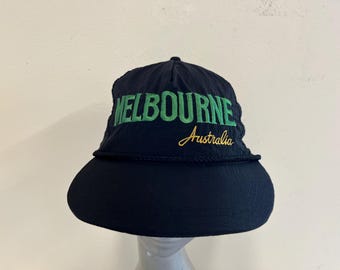 May include: A navy blue hat with a wide brim. The word "MELBOURNE" is embroidered in green across the front, with "Australia" embroidered below in gold. The hat has a rope detail around the brim.
