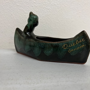 May include: A black and green ceramic canoe with a small bear figure in the front. The canoe has the words "Quebec Canada" written on the side.