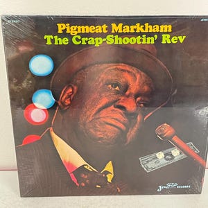 May include: A vintage vinyl record album cover featuring Pigmeat Markham. The album cover is black with a yellow title that reads "Pigmeat Markham The Crap-Shootin' Rev". The album cover features a photo of Pigmeat Markham in a black suit and a hat. He is holding a microphone and a wad of cash with dice on top. The background is a red, white, and blue color scheme.