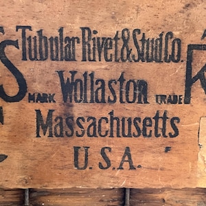 May include: Close-up of a wooden surface with black text that reads "Tubular Rivet & Stud Co. Mark Wollaston Trade Massachusetts U.S.A.". The text is surrounded by two stylized letter "R" logos.