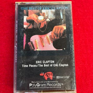May include: A vintage cassette tape of Eric Clapton's album "Time Pieces/The Best of Eric Clapton." The cover features a black background with an image of a guitar and a pocket watch. The cassette case is clear.