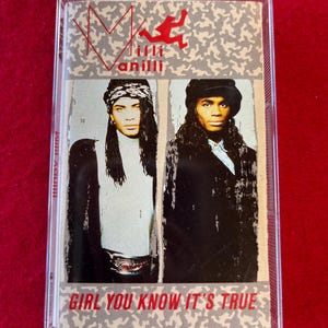 May include: A Milli Vanilli cassette tape in a clear plastic case. The cover features two people with long hair, and the text "Girl You Know It's True". The background has a gray and white pattern with the Milli Vanilli logo.