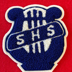 May include: A blue and white embroidered patch with the letters "SHS" in white. The patch has a shield shape with a white border and is set against a red background. The patch is likely for a school or organization.
