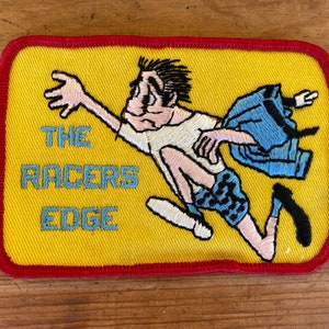 May include: Yellow embroidered patch with red stitching featuring a cartoon figure running with a blue jacket in the background. The text "THE RACERS EDGE" is embroidered in blue and white.