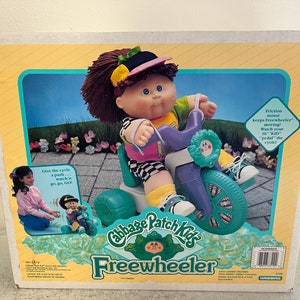 May include: A Cabbage Patch Kids Freewheeler tricycle toy with a green, purple, and blue color scheme. The tricycle has a flower-shaped handlebar and a sticker on the front wheel. The box features a Cabbage Patch Kid doll riding the tricycle.