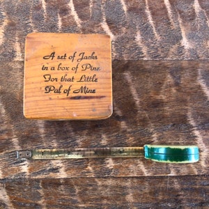 May include: A wooden box with the text "A set of Jacks in a box of Pins For that Little Pal of Mine" engraved on the top. A small, green and gold measuring tape is also pictured.