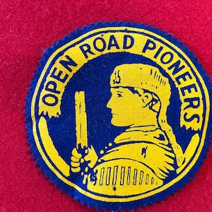 May include: A circular patch with the words "OPEN ROAD PIONEERS" in blue lettering around the edge. Inside, a yellow silhouette of a person holding a staff is featured against a blue background. The patch is on a red background.