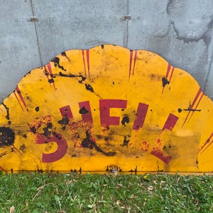 May include: A vintage, yellow and red metal sign with the word "SHELL" in red letters. The sign is shaped like a half-circle and has a worn, weathered look.