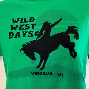 May include: Green t-shirt with a black graphic of a cowboy riding a bucking bronco under a sun. The text "WILD WEST DAYS" is above the image, and "VIROQUA, WI" is below.