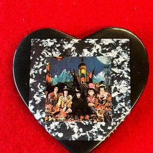 May include: A heart-shaped, black guitar pick featuring a band image. The image is set against a white and black marbled background. The pick is on a red surface.