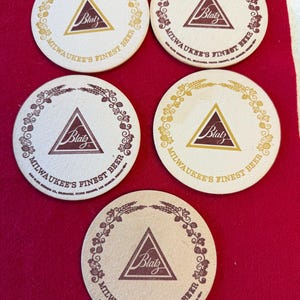 May include: Five round beer coasters with the Blatz logo in a triangle. The coasters have a cream background with a brown or gold border of wheat and the text "Milwaukee's Finest Beer".