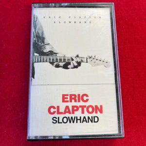 May include: A cassette tape featuring the album "Slowhand" by Eric Clapton. The cover art shows a black and white image of a person playing a guitar. The artist's name and album title are in red and black text.