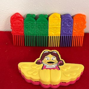 May include: A set of colorful plastic combs featuring cartoon characters. The combs are red, green, yellow, purple, and orange. There is also a separate yellow plastic toy with a cartoon character on it.