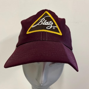 May include: A maroon baseball cap with a yellow triangle patch that says "Blatz" in white cursive lettering.