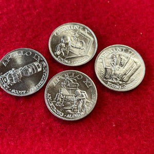 May include: Four silver-colored commemorative coins featuring images of the Apollo 11 mission, Gemini 6, and Apollo 17. The coins feature the names of the astronauts and the mission names.
