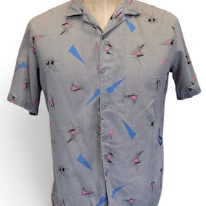 May include: A short-sleeved button-down shirt in a light gray color featuring a repeating pattern of small black geometric line clusters, solid blue triangles, and solid pink triangles across the front, collar, and sleeves.