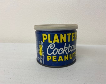 May include: A vintage Planters Cocktail Peanuts tin. The cylindrical tin is navy blue with yellow and white text. The Planters Peanut character, Mr. Peanut, is featured on the side. The lid is silver and the tin is sitting on a white surface.