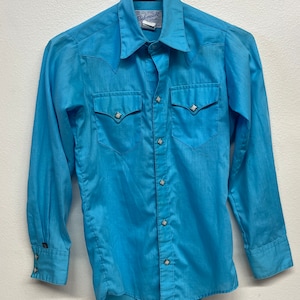 May include: A light blue Western-style shirt with long sleeves, two chest pockets, and decorative diamond-shaped accents on the pockets, cuffs, and buttons. The shirt is hanging on a black hanger.