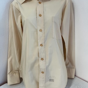 May include: A long-sleeved, light beige button-down shirt with a single chest pocket. The shirt has gold buttons and a label that reads "Sizes 14 1/2 - 15 1/2  Made in U.S.A.  Sanforized", "Rose  Reg. U.S. Pat. Off."