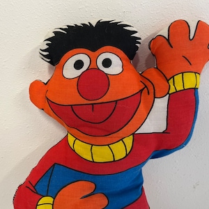 May include: A red and orange plush toy of Elmo from Sesame Street. Elmo is waving with his right arm and has a big smile on his face. Elmo is wearing a red shirt with a blue and yellow stripe.