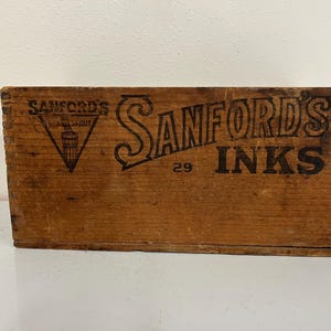 May include: A vintage wooden box with the words "SANFORD'S INKS" in black lettering. The box is rectangular and has a weathered appearance, suggesting age. The number "29" is also visible on the box.