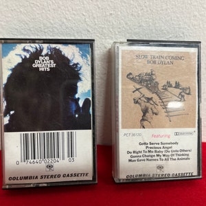 May include: Two vintage cassette tapes, one featuring Bob Dylan's Greatest Hits and the other featuring Slow Train Coming by Bob Dylan. Both tapes are black with white text and images.