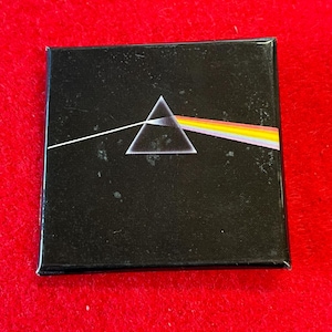 May include: Square black magnet featuring the iconic album art of a prism refracting light into a rainbow. The design includes a white triangle and a spectrum of colors. A classic music-themed item.