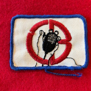 May include: Embroidered patch with a white background, a blue border, and a red circle with a microphone inside. The microphone is black, and the hands holding it are also black. The patch is rectangular.