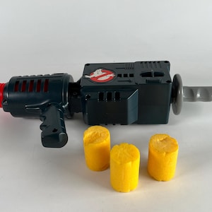 May include: A toy Ghostbusters proton pack gun in dark green and black with a red nozzle and a gray handle. The Ghostbusters logo is on the side. Three yellow foam projectiles are in front of the gun.