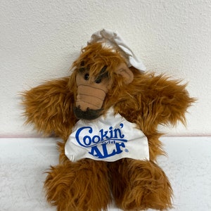 May include: A brown furry puppet wearing a white chef's hat and apron with the text "Cookin' with ALF" in blue.