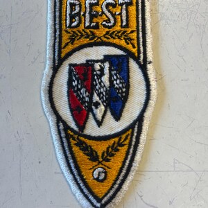 May include: Embroidered patch with the word "BEST" in white block letters above a shield design. The shield features three crests in red, black, and blue. The patch has a gold border with a white stitched edge.