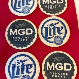 May include: Six round beer coasters with the Miller Lite and MGD Genuine Draft logos in blue and black. The coasters are arranged on a red surface. The Miller Lite coasters have a blue logo with the words "A True Pilsner Beer".