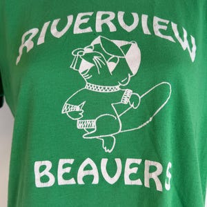 May include: Green t-shirt with the words "RIVERVIEW BEAVERS" in white block letters. A cartoon beaver mascot is in the center, wearing a hat and holding a log. The shirt is made of a soft material.