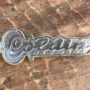 May include: A silver metal key-shaped emblem with the word "Coldin" and "NEW YORK" in raised lettering.