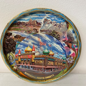 May include: A vintage metal tray with a colorful illustration of the World's Only Corn Palace in Mitchell, South Dakota. The tray also features images of Mount Rushmore, Badlands National Monument, a bison, a ring-necked pheasant, and a prairie flower.