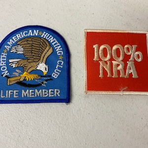 May include: Two embroidered patches. The first is blue with a white eagle and the text "North American Hunting Club Life Member". The second is orange with white text that reads "100% NRA".