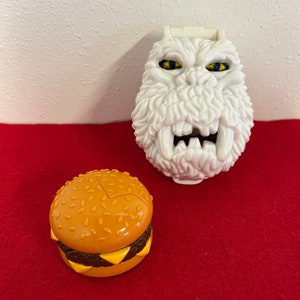 May include: Two plastic toys, a white monster with yellow eyes and a yellow and brown hamburger.